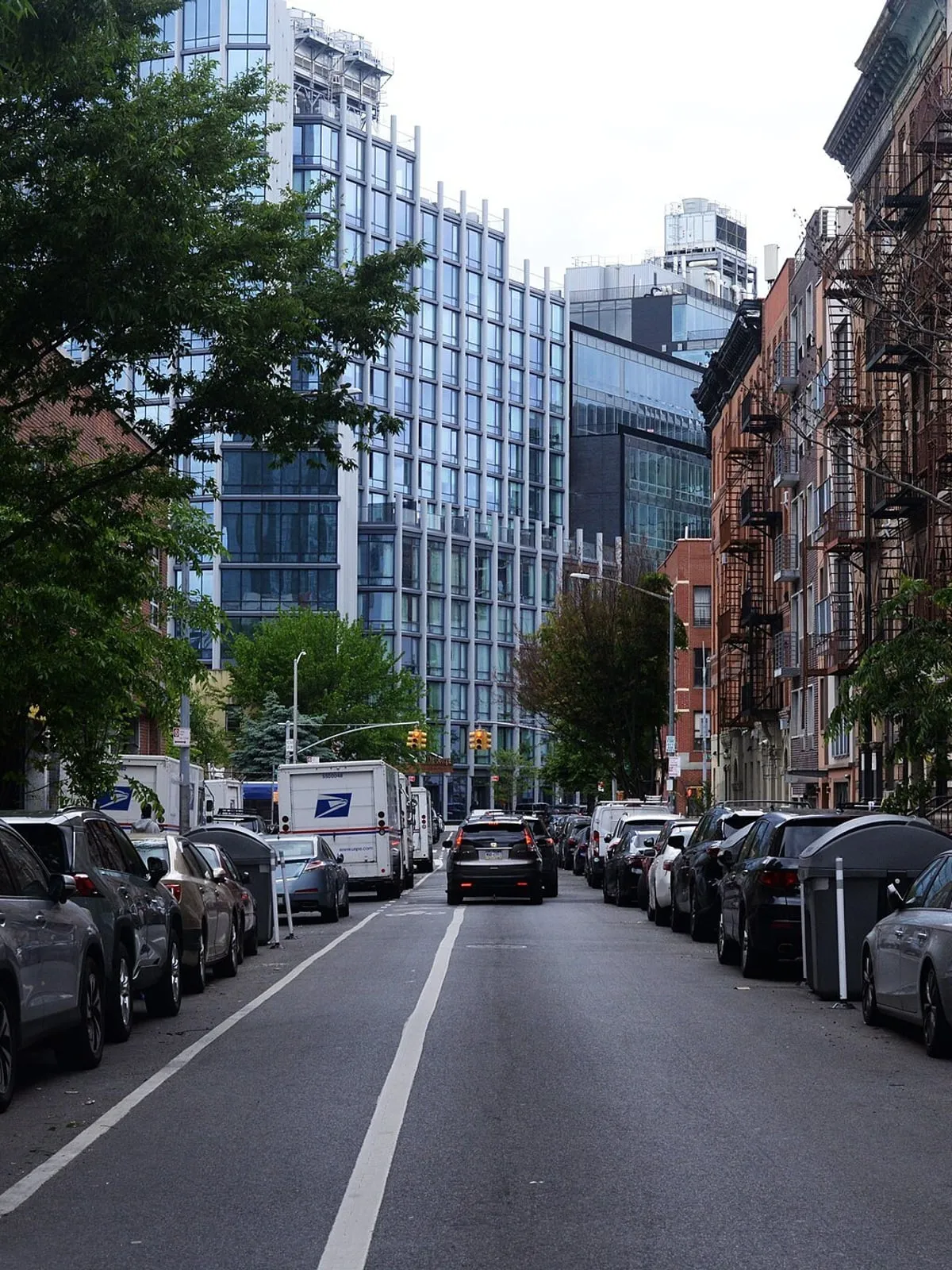 Row of walk-up buildings along a Manhattan side street, fire escapes over the storefronts and cars parked either side.