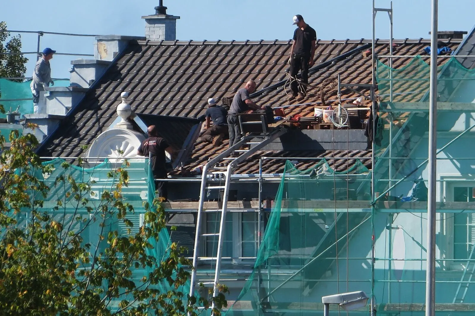 Roofing crew working across a stripped roof slope, the building wrapped in scaffold and netting below them.