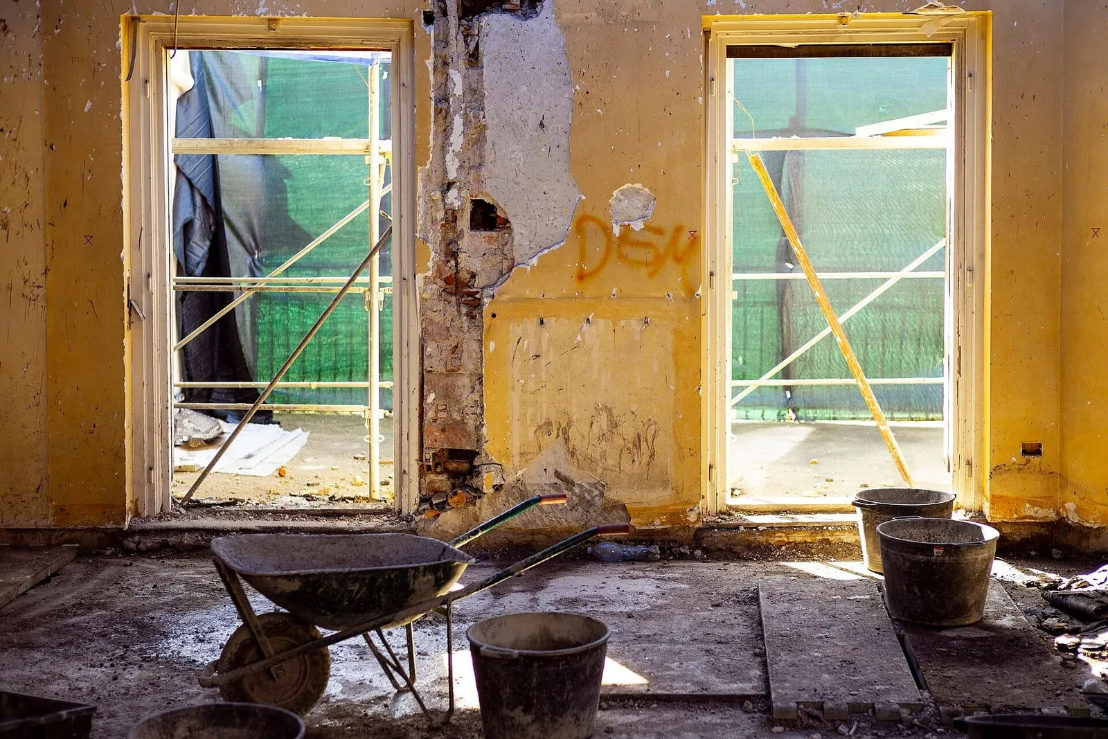 Gutted room mid-reconstruction, plaster stripped back past the window reveals, wheelbarrow and buckets on the bare floor.