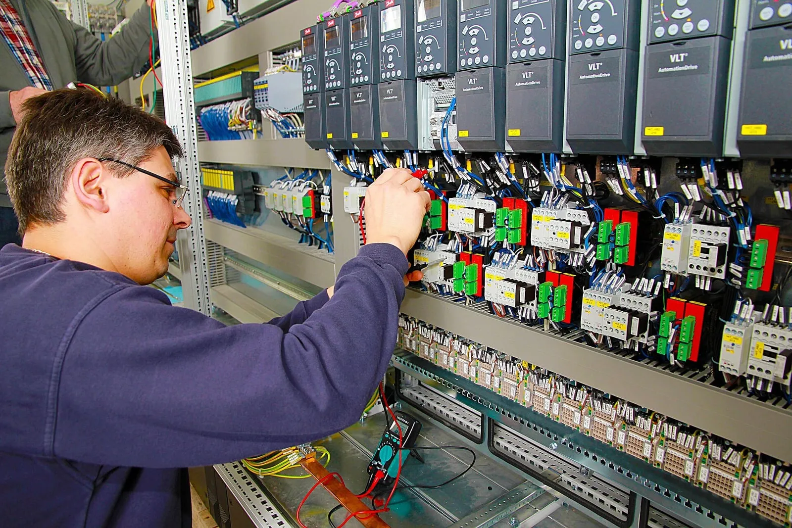 Hand at an open building control panel, variable-speed drives and wired terminal blocks racked above.