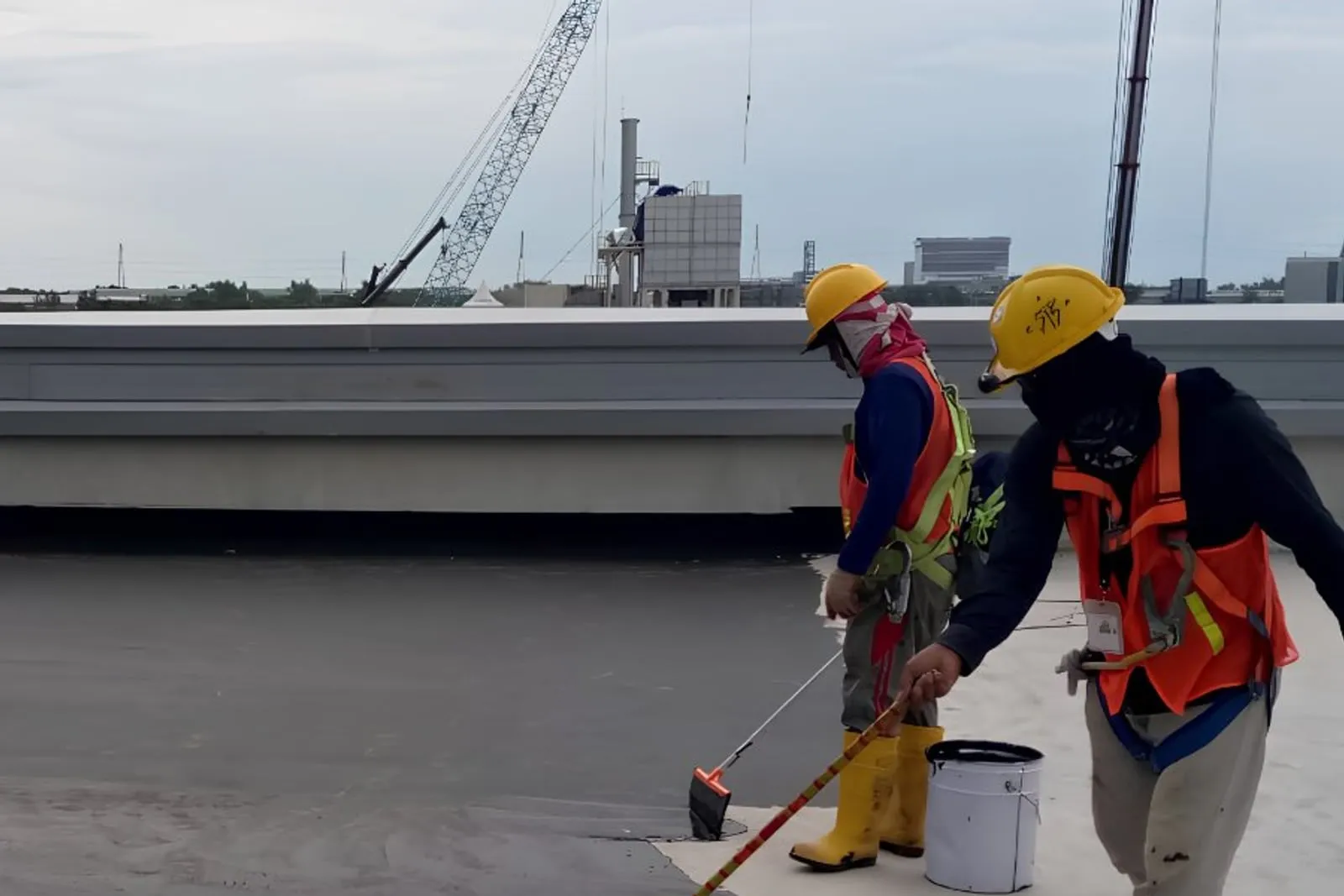 Two workers in hi-vis spreading waterproofing membrane across a flat roof deck, tower cranes on the skyline behind them.